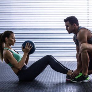 A woman doing sit-ups while holding a med ball and being assisted by her partner.