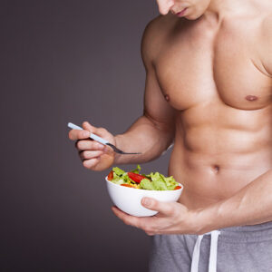 A fit man showing his abs while eating a vegetable salad.