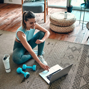 A fit woman sitting on the carpet at home while on her laptop with a tumbler and dumbbells on her side.