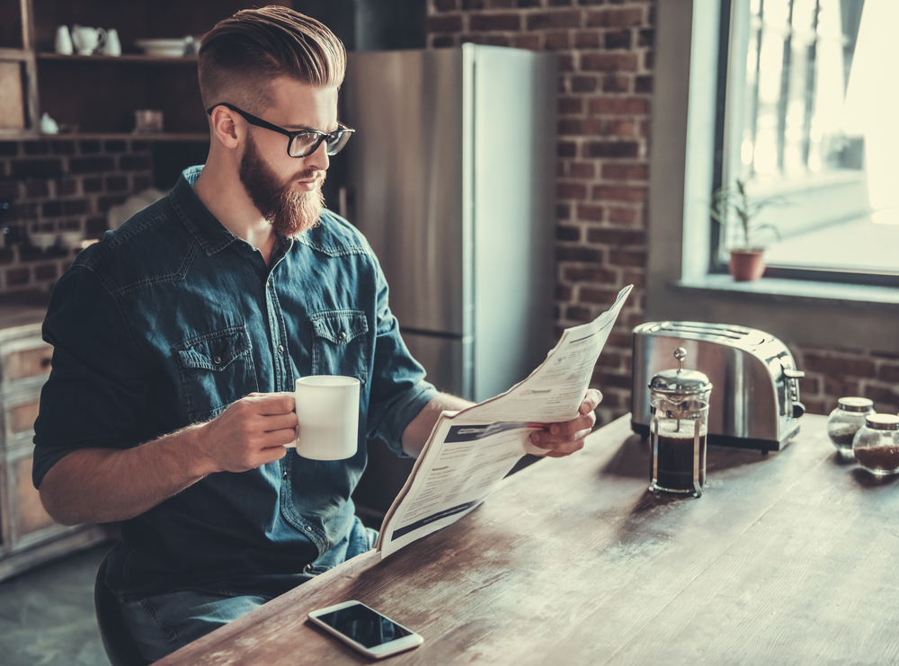 Young man is drinking coffee and reading a newspaper while resting in kitchen