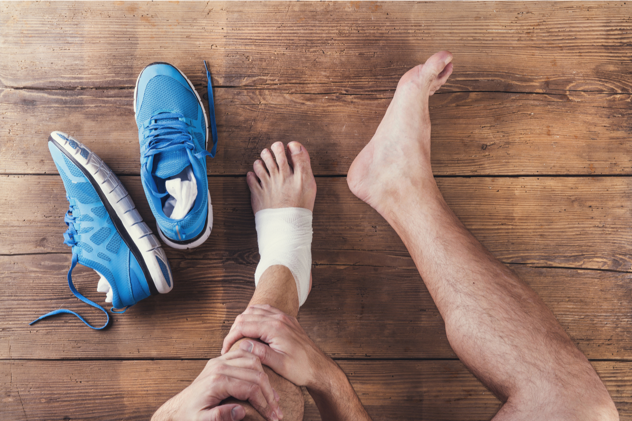 A man sitting on a wooden floor with bandage on his left foot because it was sprained and a pair of running shoes on the side. He is thinking what is good to treat sprain.