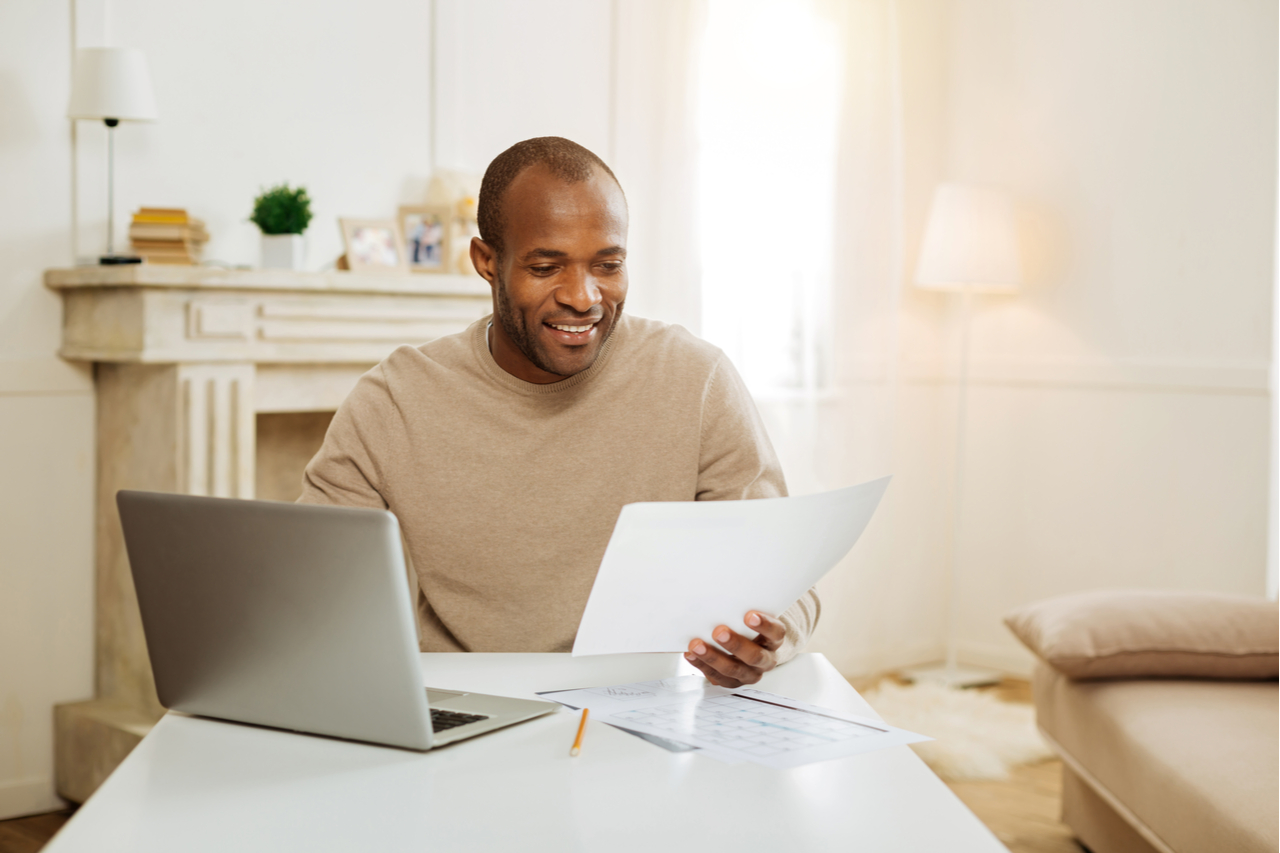 An Afro-American man in front of his laptop, holding some papers on his hand while working from home - one of his financial resources. 