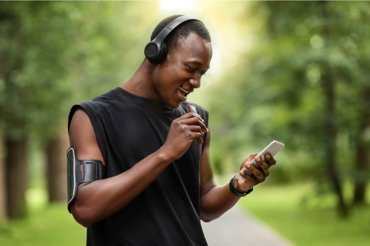 Cheerful african sportsman eating protein bar and using smartphone while resting.