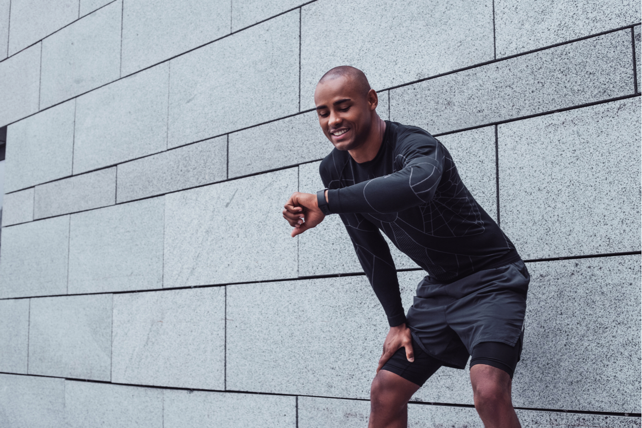 Young man in sportswear looking at his smartwatch with smile while standing against grey background outdoors.