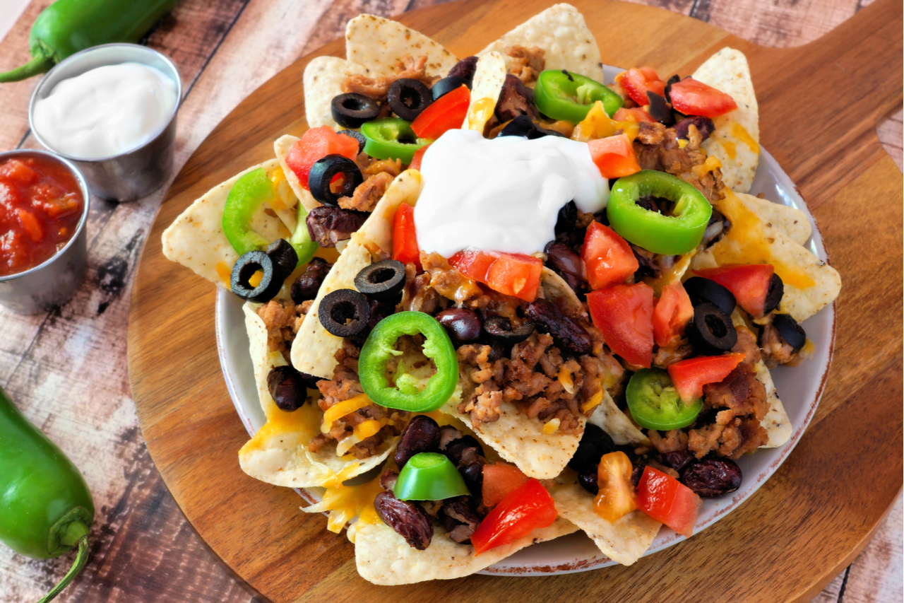 Nacho chips topped with sour cream, ground meat, jalapenos, tomatoes, beans and melted cheese on a wooden paddle board.