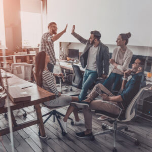 We did it! Two cheerful young business people giving high-five while their colleagues looking at them and smiling