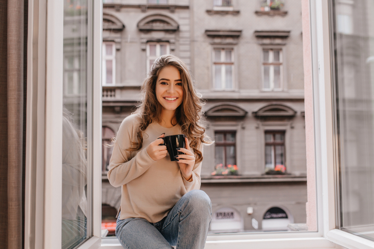 Adorable woman expressing positive emotions while posing on sill with cup of golden milk latte.