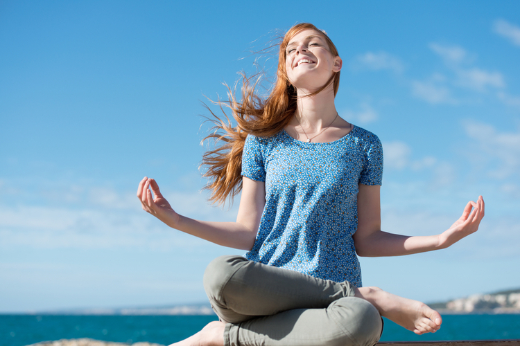 Beautiful woman meditating at the seaside sitting cross-legged on the rocks with her head tilted to the sun and her eyes closed. COVID weight gain is it normal?