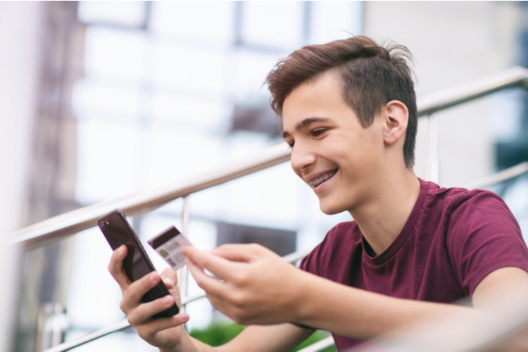 Teenage boy with greenlight debit card and mobile phone makes purchasing outdoors.