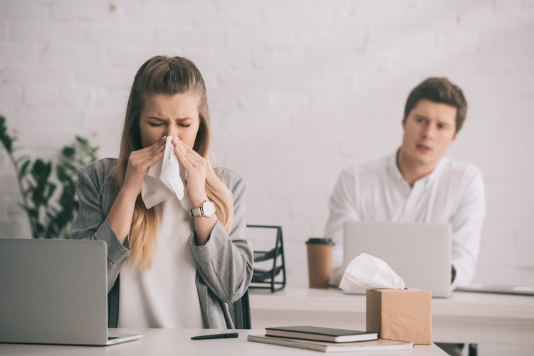 Businesswoman sneezing in tissue near coworker in office reacting to workplace stress.