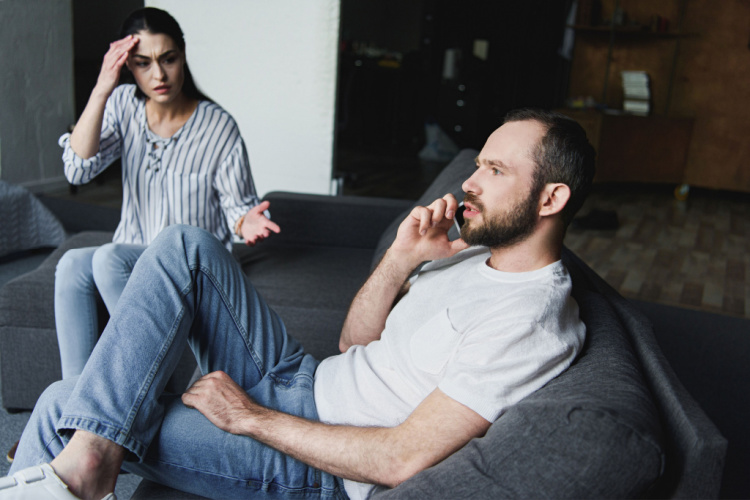 upset woman talking to her husband while he talking by phone on couch