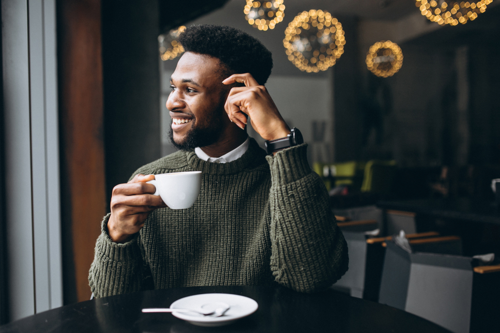 Man at a cafe drinking a cup of coffee, looking out the window and smiling 