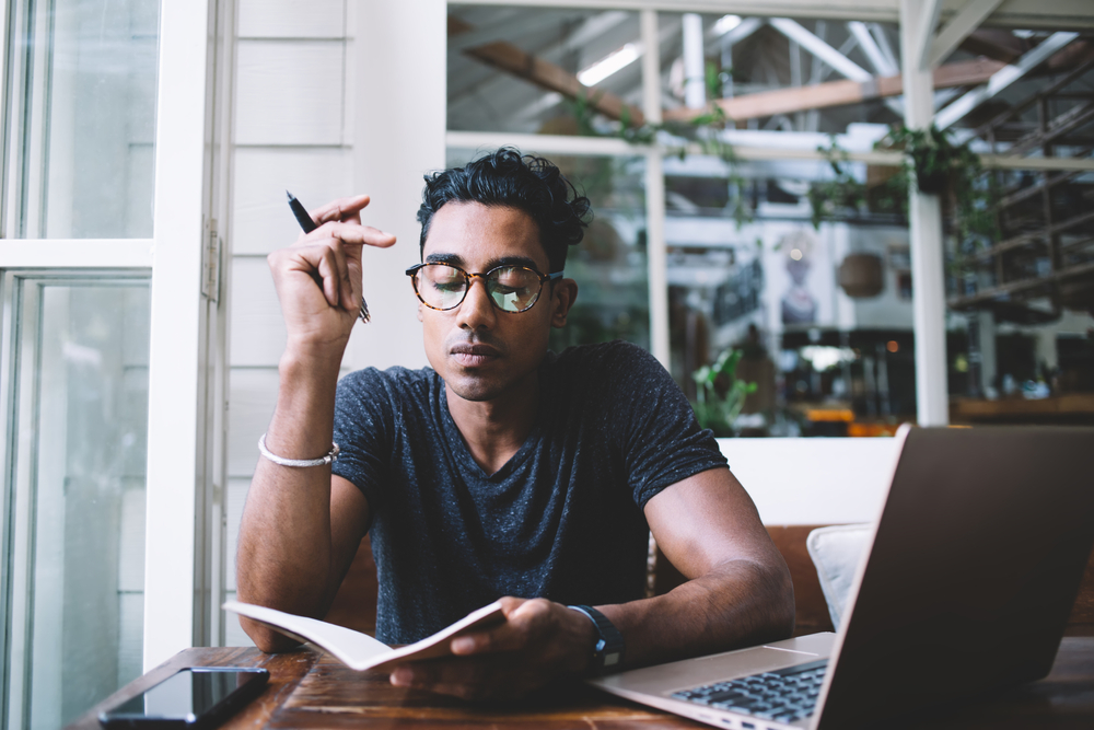 man thinking and taking notes while sitting at table with laptop